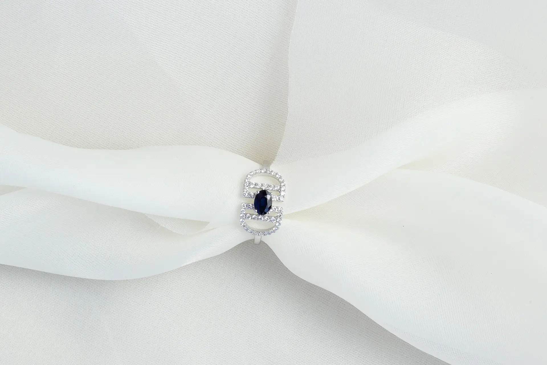 A close-up of a ring featuring a dark blue oval gemstone surrounded by smaller clear stones on a white metal band, displayed on a softly folded white fabric.